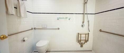A small, white-tiled bathroom with a toilet, a shower, and a handrail.
