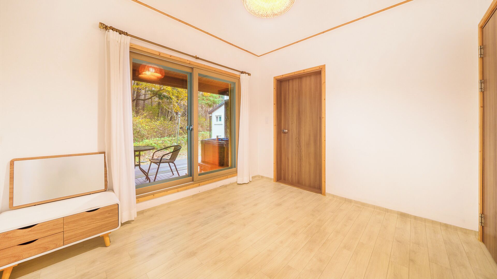 A room with a wooden floor, a sliding glass door leading to an outdoor area with a table and chairs, and a wooden door.