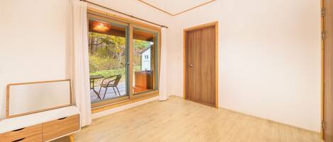 A room with a wooden floor, a sliding glass door leading to an outdoor area with a table and chairs, and a wooden door.