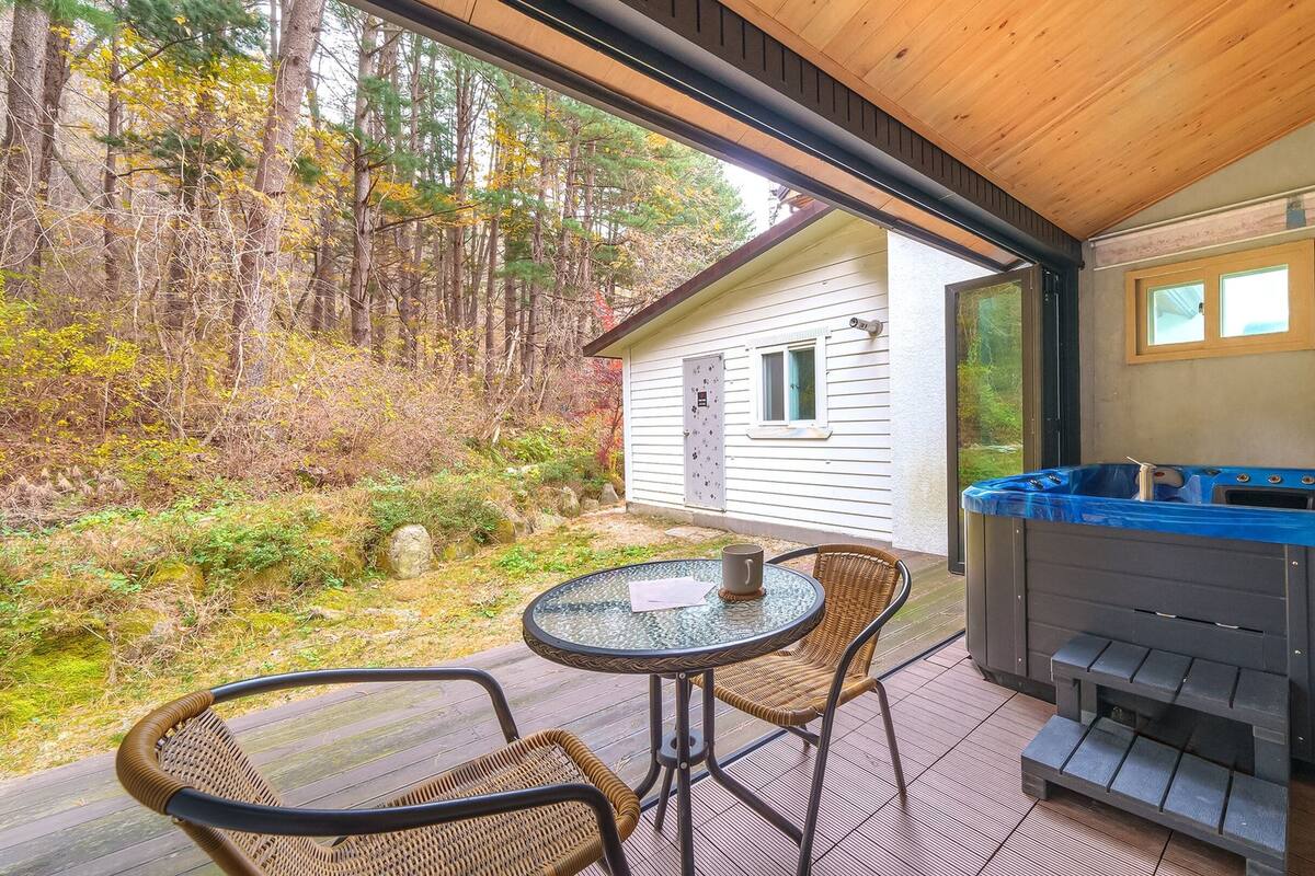 Überdachte Terrasse mit Whirlpool, Korbstühlen und Blick auf eine Hütte im Wald.