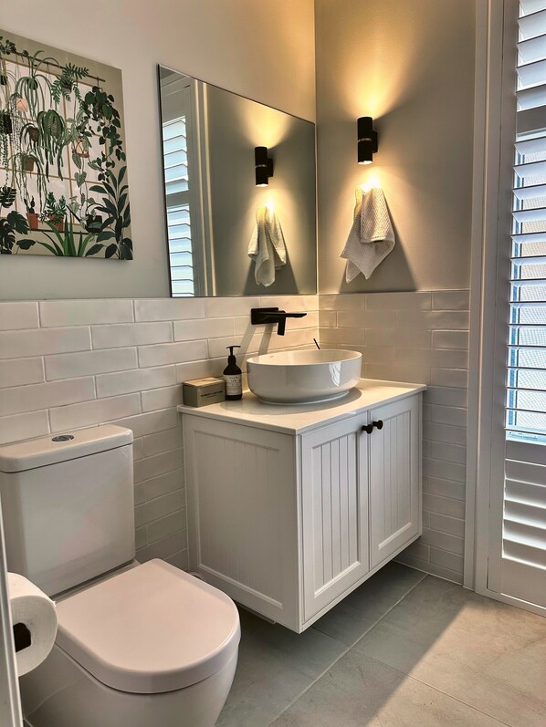 A modern bathroom with a white toilet, a white sink with a vessel basin, and a mirror above the sink. There are two wall-mounted lights above the mirror, and a towel rack with a towel hanging on it. - Luxury Home for Work Teams and Families (Muswellbrook)