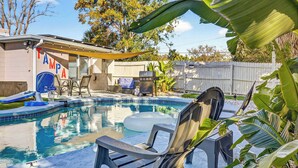A backyard with a swimming pool, lounge chairs, a barbecue grill, and a covered patio area.