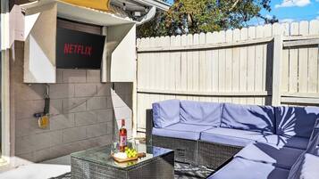 An outdoor seating area with a Netflix sign, a glass table with a bottle and fruit, and a white picket fence.