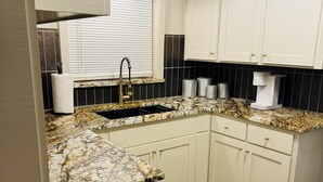 A kitchen with white cabinets, a granite countertop, a stainless steel sink, a black trash bin, and a window with blinds.