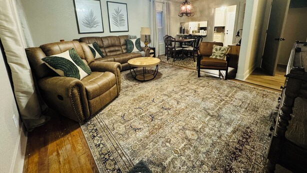 A living room with a brown leather sofa, a round coffee table, and a patterned rug. There is a ceiling fan, a chandelier, and a dining area in the background.