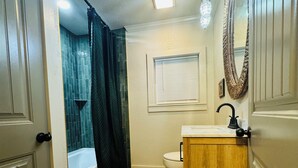 A bathroom with a wooden vanity, a white toilet, a shower with a dark curtain, and a window with blinds.