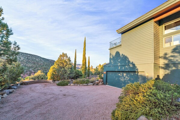 A house with a gravel driveway, surrounded by various shrubs and trees, and a mountainous backdrop.