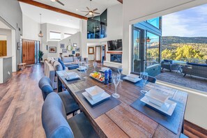 A spacious dining area with a long wooden table set for four, a large window offering a view of the outdoors, and a modern kitchen in the background.