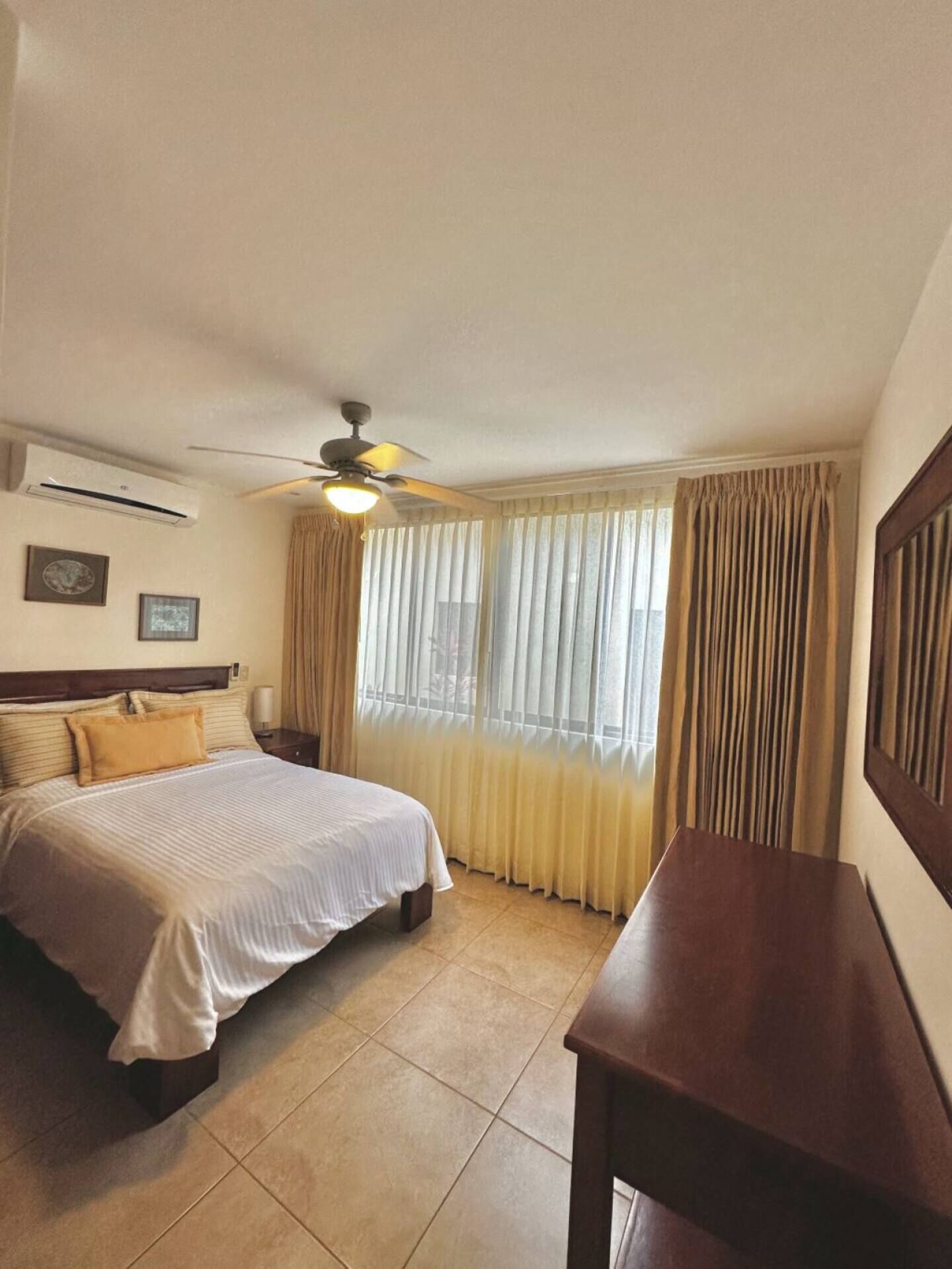 Quarto de hotel com cama, mesa de cabeceira, ventoinha no teto, janela com cortinas e uma mesa de madeira.