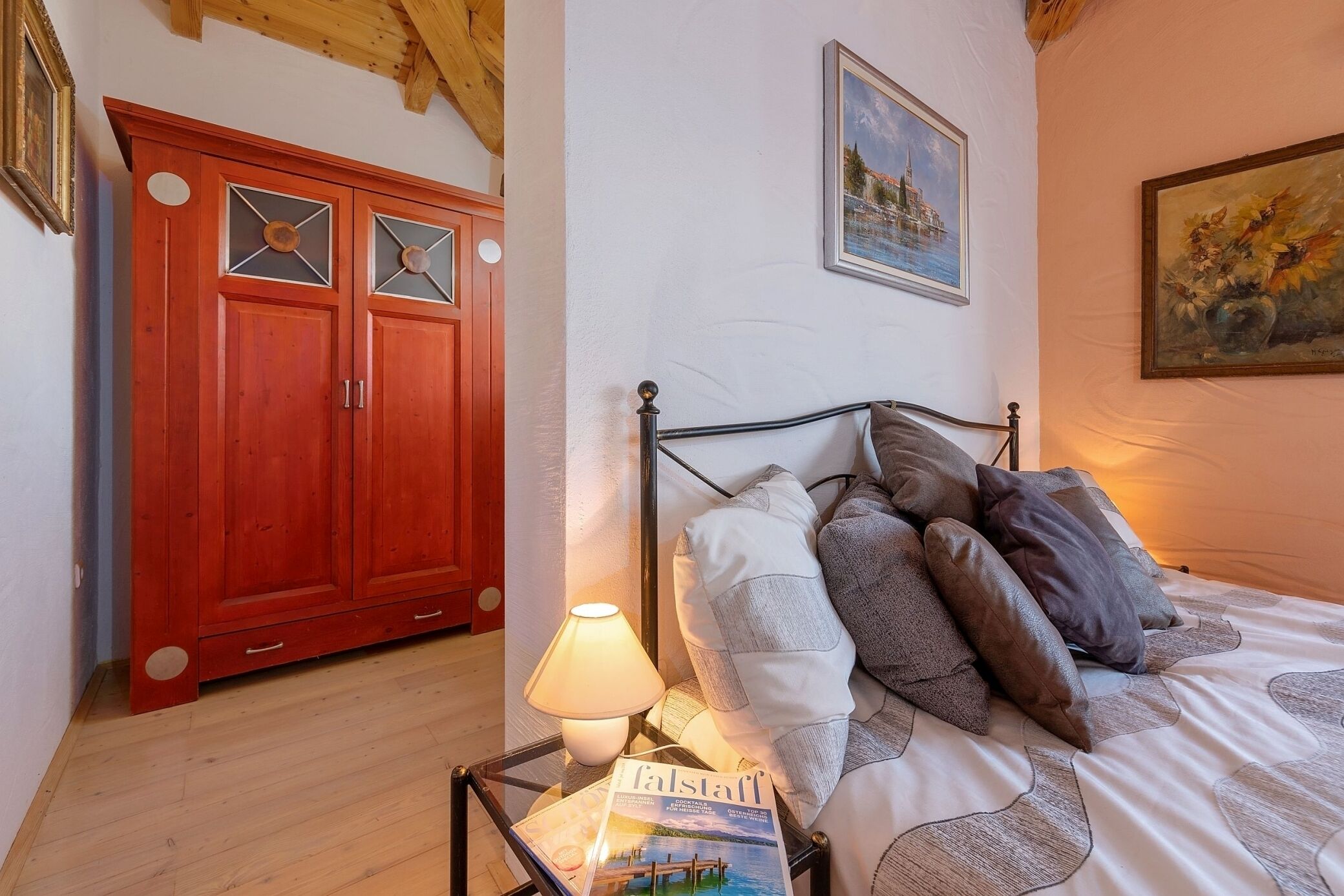 Un dormitorio con una cama, almohadas, una revista, una lámpara y un armario rojo.