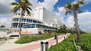 A modern hotel with a curved facade, palm trees, and a pathway leading to the entrance. - Castle 1133 Ocean view studio, Pool and Beach Access, FREE Parking, Gym, Tennis and more! (Miami Beach)