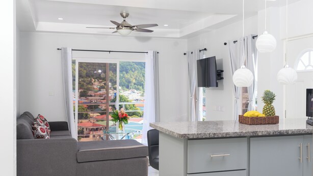A modern kitchen with a granite countertop, grey cabinets, and a dining area with a grey sofa and a small table. - Caldera Villas Where Nature Meets Comfort (Soufriere)