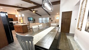 A spacious dining area with a long wooden table and white chairs, a kitchen with wooden cabinets and a black refrigerator, and a ceiling fan.