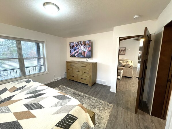 A bedroom with a bed, a dresser, a TV mounted on the wall, and a window with blinds.