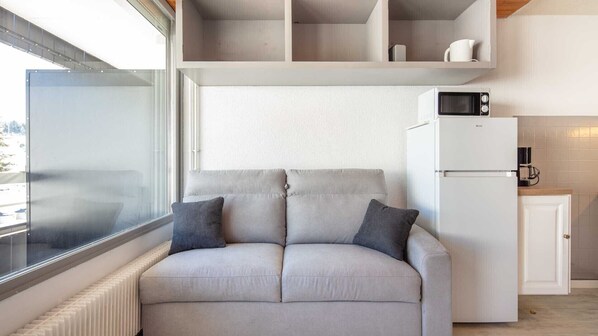 A modern living room with a sofa, a radiator, a window, and a kitchen area with a refrigerator and microwave.