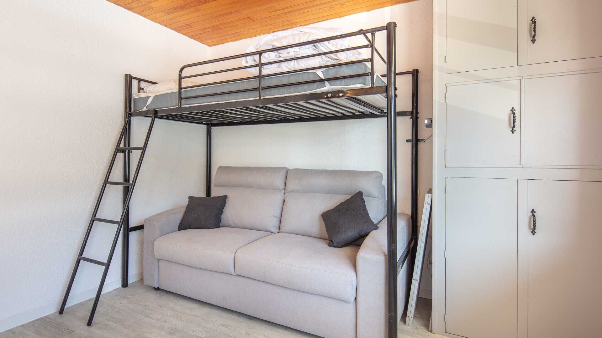 A bunk bed with a ladder, a sofa, and a wardrobe in a room with wooden ceiling beams.