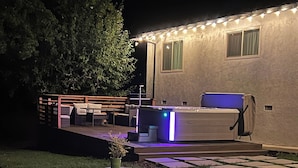 Un área de piscina con hidromasaje y asientos en terraza, rodeada de una terraza de madera y una casa con luces colgantes.