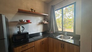 A compact kitchen with a black countertop, wooden cabinets, a built-in microwave, a kettle, and a window with a view of trees. - Tulum Ayook apartment with Starlink (Tulum)