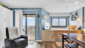 Une pièce compacte avec une porte-fenêtre coulissante donnant sur un balcon, un lit avec une couverture à rayures, un bureau avec une lampe et une chaise.