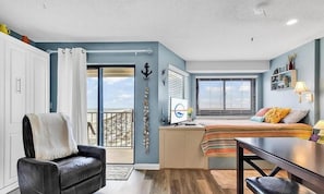 Una habitación compacta con puerta corrediza de vidrio a un balcón, una cama con manta a rayas, un escritorio con lámpara y una silla.
