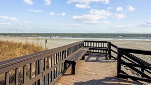 Une promenade en bois menant à une plage, avec un ciel dégagé et l’océan en arrière-plan.