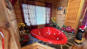 A wooden cabin interior with a red bathtub, stone flooring, wooden walls, a window with curtains, and various decorations including a teddy bear and a Christmas truck. - Honeymoon log cabin with heart shaped whirlpool Tub & Hot Tub on Covered Porch (Sevierville)