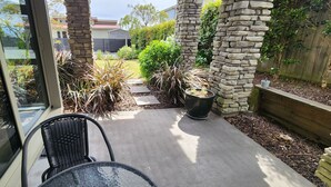 A patio area with a glass door, a black wicker chair, and a table. There are stone pillars and a small garden with various plants. - CENTRAL RETREAT Spa Bath, short walk to 
Lake/town center, Sunsets & some views. (Taupō)