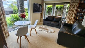 A living room with a black leather sofa, a white chair, a small round table with a vase of flowers, and a bookshelf. - CENTRAL RETREAT Spa Bath, short walk to 
Lake/town center, Sunsets & some views. (Taupō)