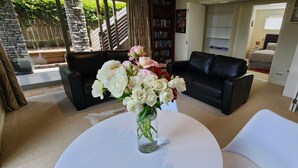 A living room with a white table, black leather sofas, a bookshelf, and a view of a garden through a glass door. - CENTRAL RETREAT Spa Bath, short walk to 
Lake/town center, Sunsets & some views. (Taupō)