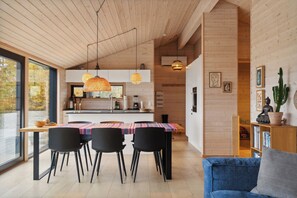 A modern kitchen with wooden cabinets, a dining table with chairs, pendant lighting, and a bookshelf with books and a cactus.