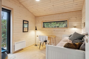 Et interiør af en træhytte med en seng, et lille bord, en stol, en radiator og et vindue med udsigt over grøn vegetation.