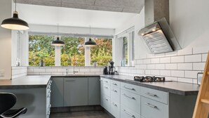 Een moderne keuken met een groot eiland, hangende lampen, een ingebouwde kookplaat en uitzicht op bomen door het raam.