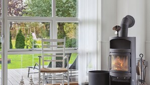 Een kamer met een groot raam, een witte schommelstoel, een houtkachel en uitzicht op een tuin.