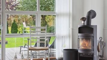 A room with a large window, a white rocking chair, a wood-burning stove, and a view of a garden.