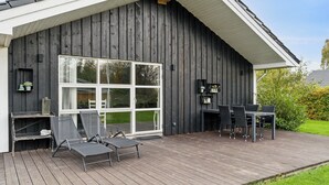 Een houten terras met tuinmeubilair, een groot raam en een zwarte buitenmuur.