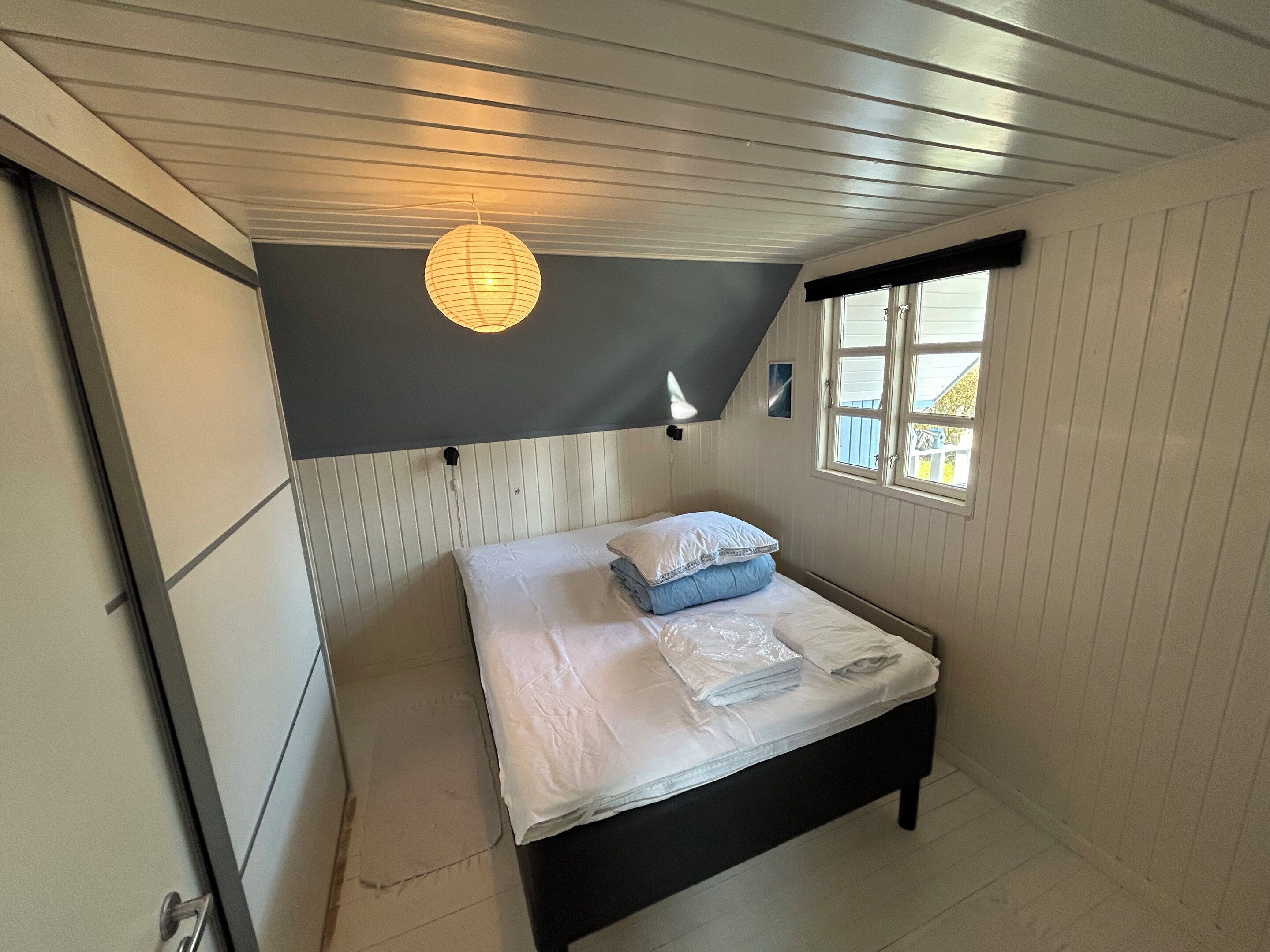 A bedroom with a bed, a window, a wall-mounted light fixture, and a door.