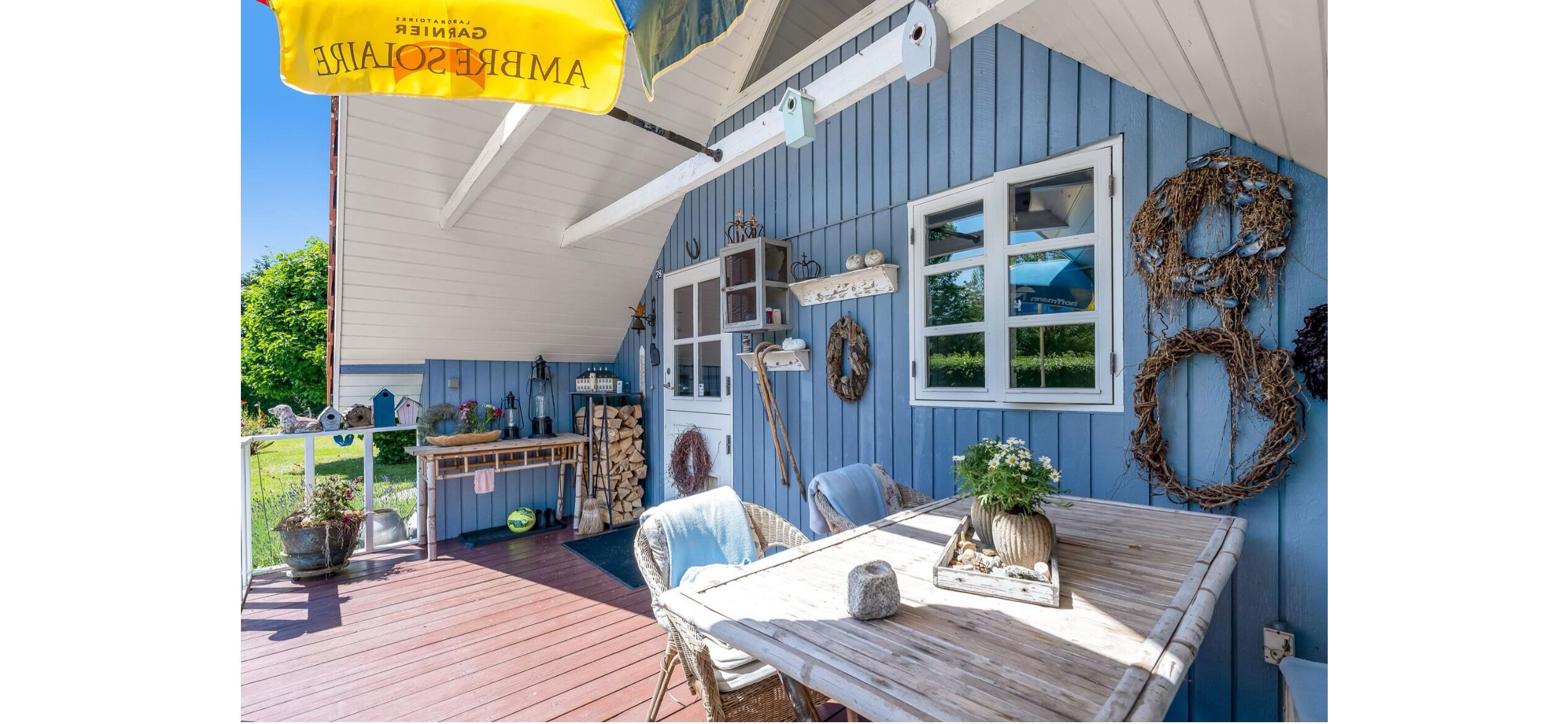 Un bâtiment bleu avec une terrasse en bois, une table ornée d’une plante et un drapeau jaune suspendu.