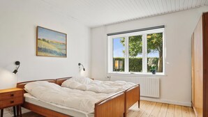 Un dormitorio con cama de madera, una mesita de noche, una lámpara, un ventanal con vistas al verdor y un cuadro en la pared.