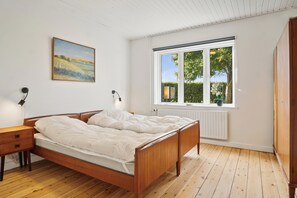 A bedroom with a wooden bed, a bedside table, a lamp, a window with a view of greenery, and a painting on the wall.
