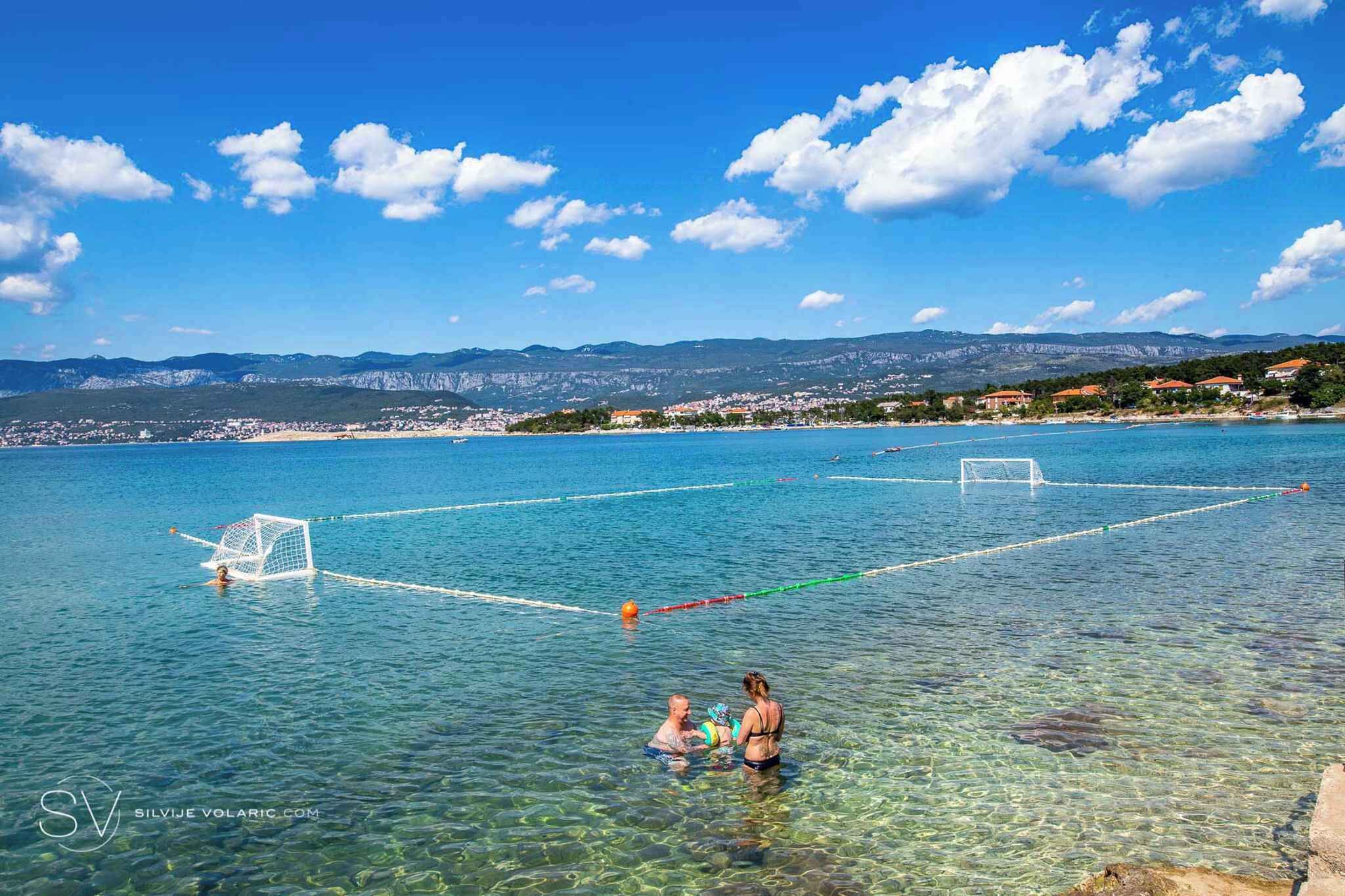Ein klarer Blick auf ein Beachvolleyballfeld mit zwei Spielern und Netz, umgeben von klarem Wasser und fernen Bergen.
