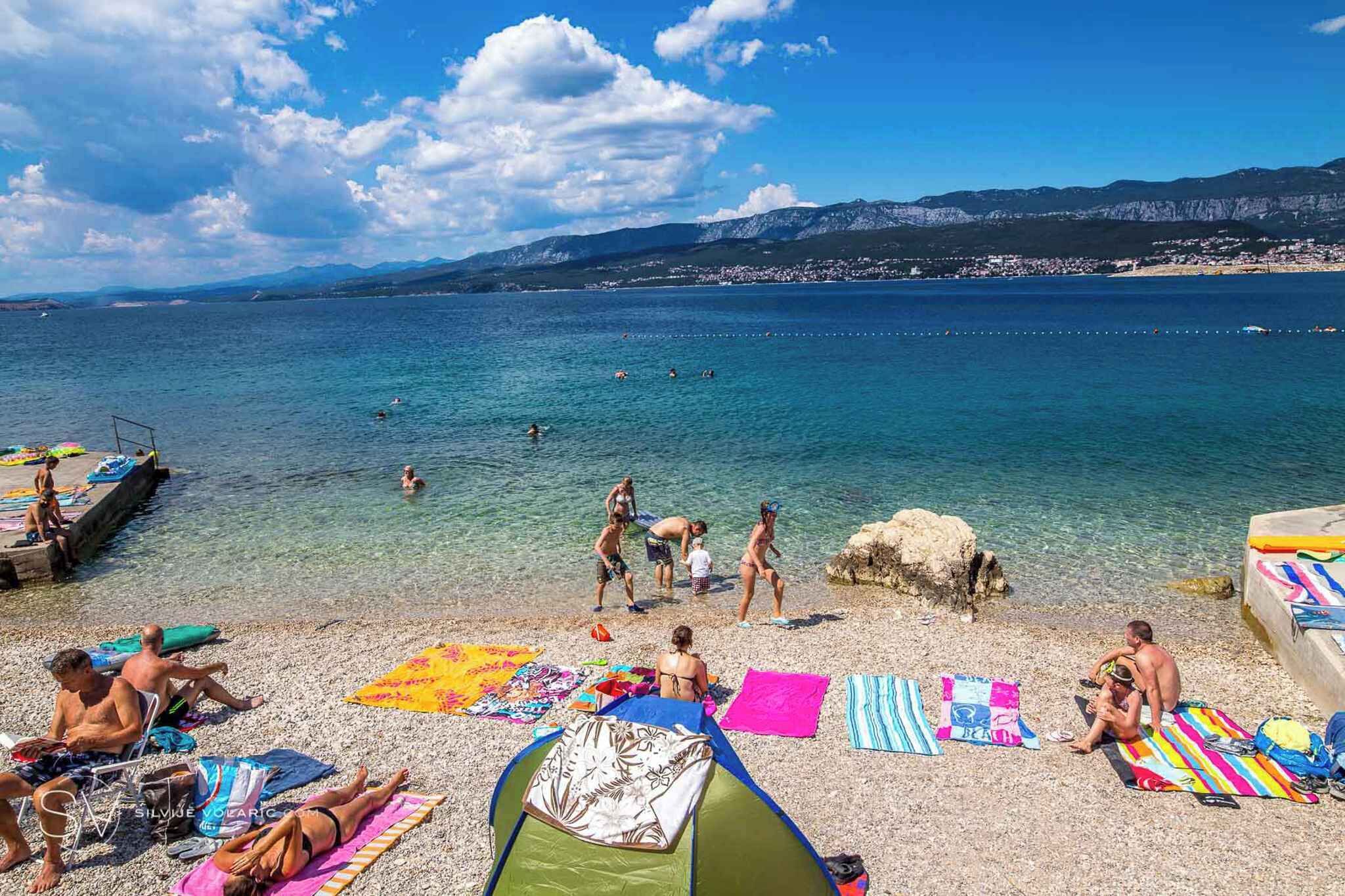 Ein Strand mit badenden und sonnenden Personen, einem Zelt und einer felsigen Küstenlinie.