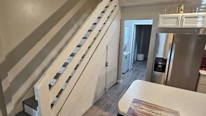 Un escalier avec des rampes blanches menant à un palier, un coin cuisine avec un réfrigérateur en acier inoxydable et une table à manger avec un set de table.