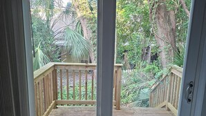 On aperçoit, à travers une porte vitrée, une terrasse en bois dotée d’une balustrade, le tout étant entouré d’une végétation luxuriante.