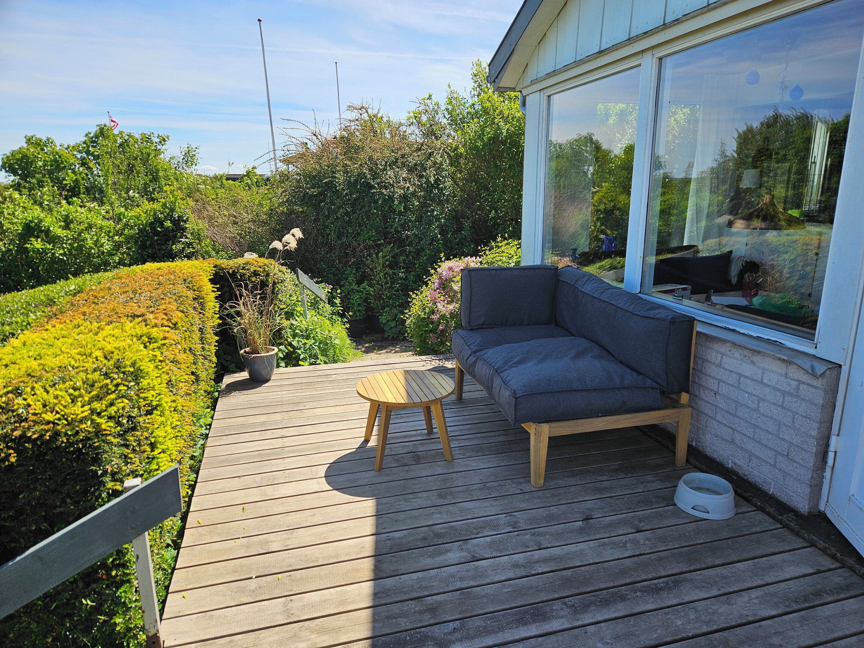 Terraza de madera con sofá y mesa, rodeada de follaje y un edificio con amplios ventanales.