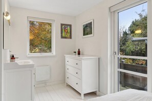 Une salle de bain avec un lavabo blanc, une fenêtre donnant sur des arbres, un meuble blanc avec des tiroirs et une porte menant à un espace extérieur.