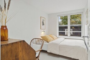 A bedroom with a bed, a chair, a desk, a vase with dried plants, a framed picture on the wall, and a window with a view of trees.