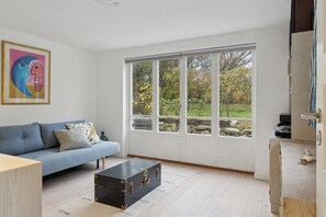 A room with a blue sofa, a wooden coffee table, a large window with a view of trees, and a painting on the wall.