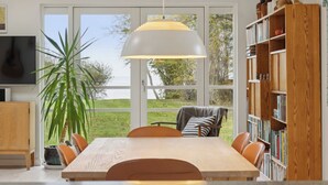 Un espace repas comprenant une table, des chaises, un luminaire suspendu, une bibliothèque et une vue dégagée sur l’extérieur grâce à de grandes fenêtres.