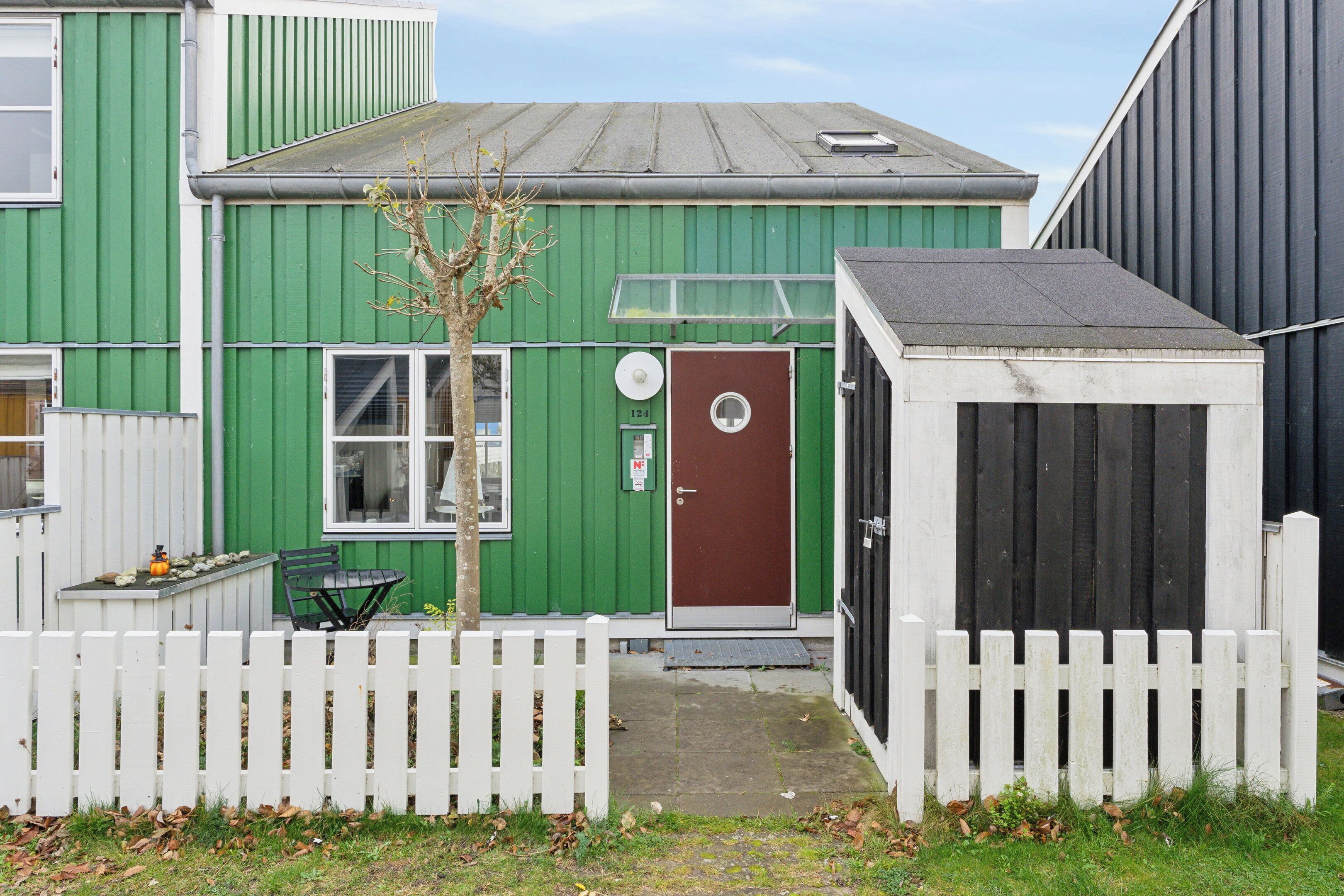 A green building with a brown door, a white picket fence, and a small tree.