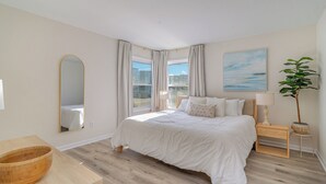 A bedroom with a large bed, a wooden bedside table, a mirror, a window with curtains, a plant, and a painting on the wall. - Steps to Beach! Spacious 3bd with pool! Murrells Inlet! Family friendly (Murrells Inlet)
